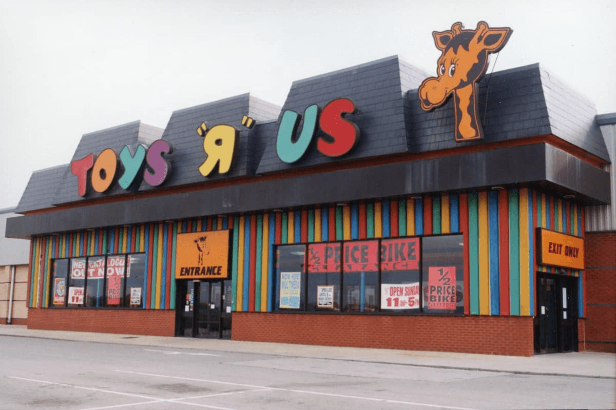The iconic brown-shingled exterior storefront of a standalone 1990s Toys "R" Us, complete with rainbow-striped wall panels and the retro logo starring Geoffrey the Giraffe on the roof.