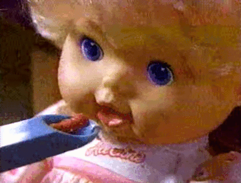 An animated GIF from a vintage Baby Alive TV commercial showing a close-up of the mechanical doll opening her mouth to accept a blue spoon of toy food.