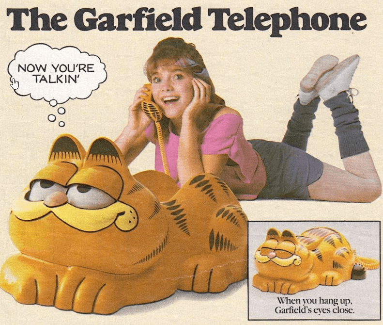 A print advertisement for 'The Garfield Telephone' showing a girl lying down using a novelty orange Garfield cat landline phone, with an inset image demonstrating how Garfield's eyes close when the receiver is hung up.