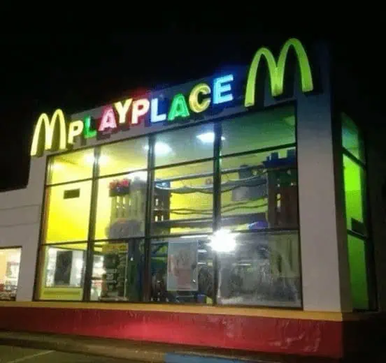 A glowing exterior night shot of a 1990s McDonald's featuring the illuminated green 'PlayPlace' signage over multi-story glass windows housing indoor climbing tubes and ball pits.