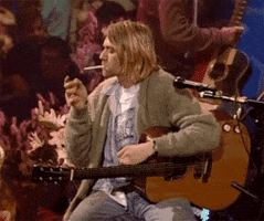 An animated GIF from Nirvana's iconic 1993 MTV Unplugged session, showing Kurt Cobain in his famous green cardigan holding a cigarette while tuning his acoustic guitar. - click to watch