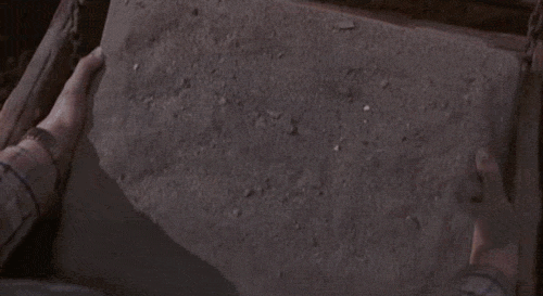 An animated GIF from Jumanji showing young Alan Parrish brushing dust off a heavy stone board game buried in dirt, revealing the mysterious, hand-carved 'JUMANJI' title header.