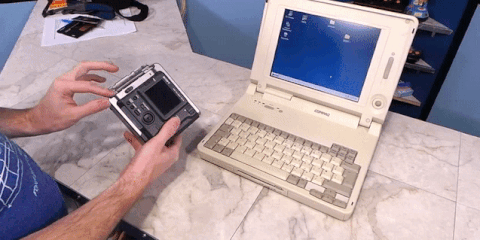 hands ejecting a 3.5-inch floppy disk directly out of the side slot of a chunky Sony Digital Mavica camera next to a vintage Compaq laptop.