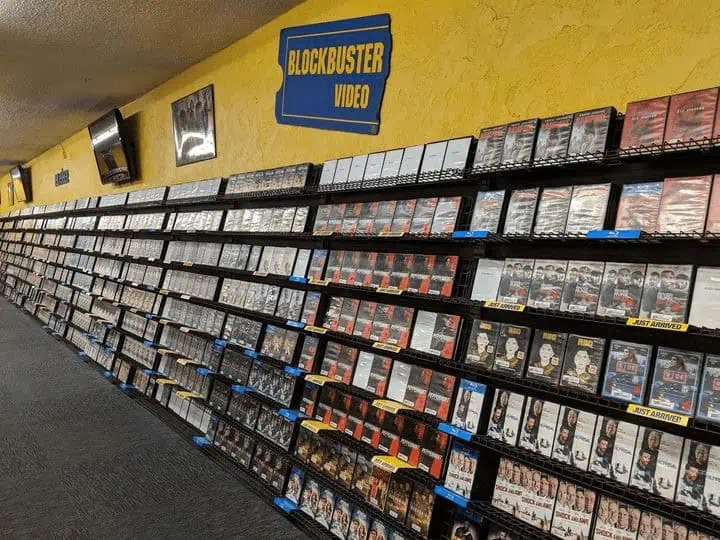 A view down a brightly lit aisle inside a 1990s Blockbuster Video, showing wall-to-wall movie rentals beneath a bold yellow-and-blue Blockbuster sign.