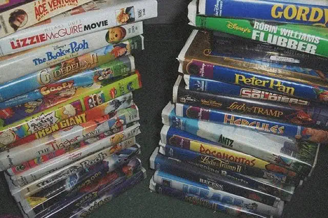 A tall stack of white plastic 90s and early-2000s VHS clamshell cases, including Ace Ventura: When Nature Calls, The Lizzie McGuire Movie, A Goofy Movie, The Rugrats Movie, and The Iron Giant.