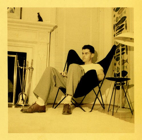 A vintage yellowed photograph of a man relaxing in a black canvas BKF butterfly chair inside a mid-century modern living room next to a fireplace and retro tripod table.