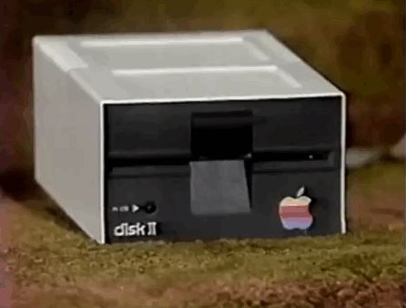 An animated GIF showing a classic beige-and-black Apple Disk II 5.25-inch external floppy drive sitting on a grassy surface with its rainbow logo.
