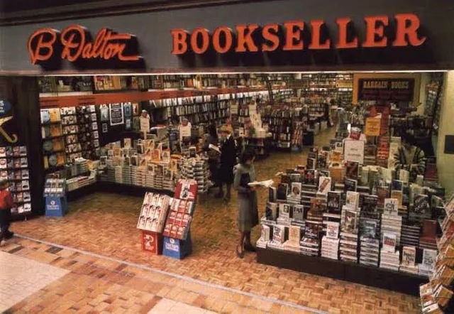 The inviting storefront of a B. Dalton Bookseller inside a carpeted 90s mall, displaying floor-to-ceiling stacks of paperbacks, bestseller tables, and cozy reading nooks.