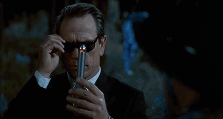 An animated GIF from Men in Black featuring Agent K (Tommy Lee Jones) sliding on his Ray-Bans and raising his shiny metallic Neuralyzer, moments before triggering the blinding flash that erases civilian memories.