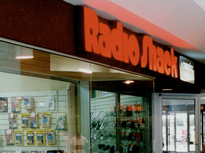 A classic mall corridor view of a RadioShack storefront, showing glass display cases packed with electronic components, handheld radios, and RC cars.