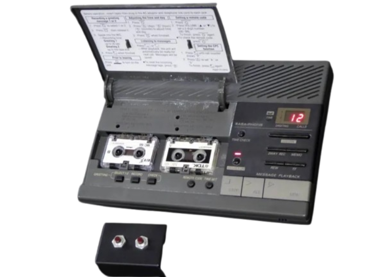 A vintage dark-grey dual-cassette telephone answering machine with an open top lid showing two microcassettes, physical playback control keys, and a bright red 2-digit LED call counter.