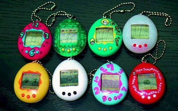 A static photo displaying eight different 1990s Bandai Tamagotchi virtual pet egg keychains lined up on a dark surface.