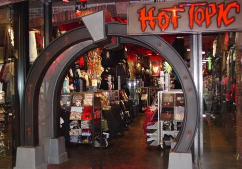 A classic 90s Hot Topic storefront featuring its signature dark industrial metal archway entrance, gothic font marquee, and racks packed with band tees, fishnets, and studded belts.