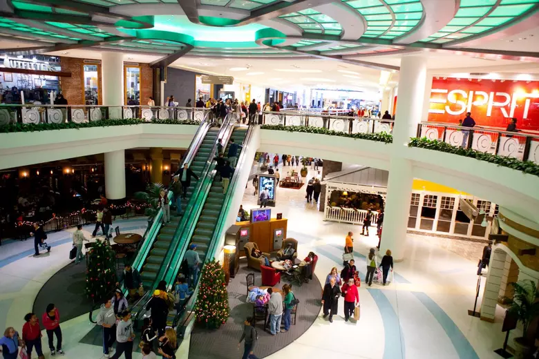 A bustling 1990s two-story shopping mall atrium lit by glowing neon green skylight ceiling panels, featuring glass escalators, shoppers, decorated Christmas trees, an Urban Outfitters, and a massive illuminated Esprit storefront.