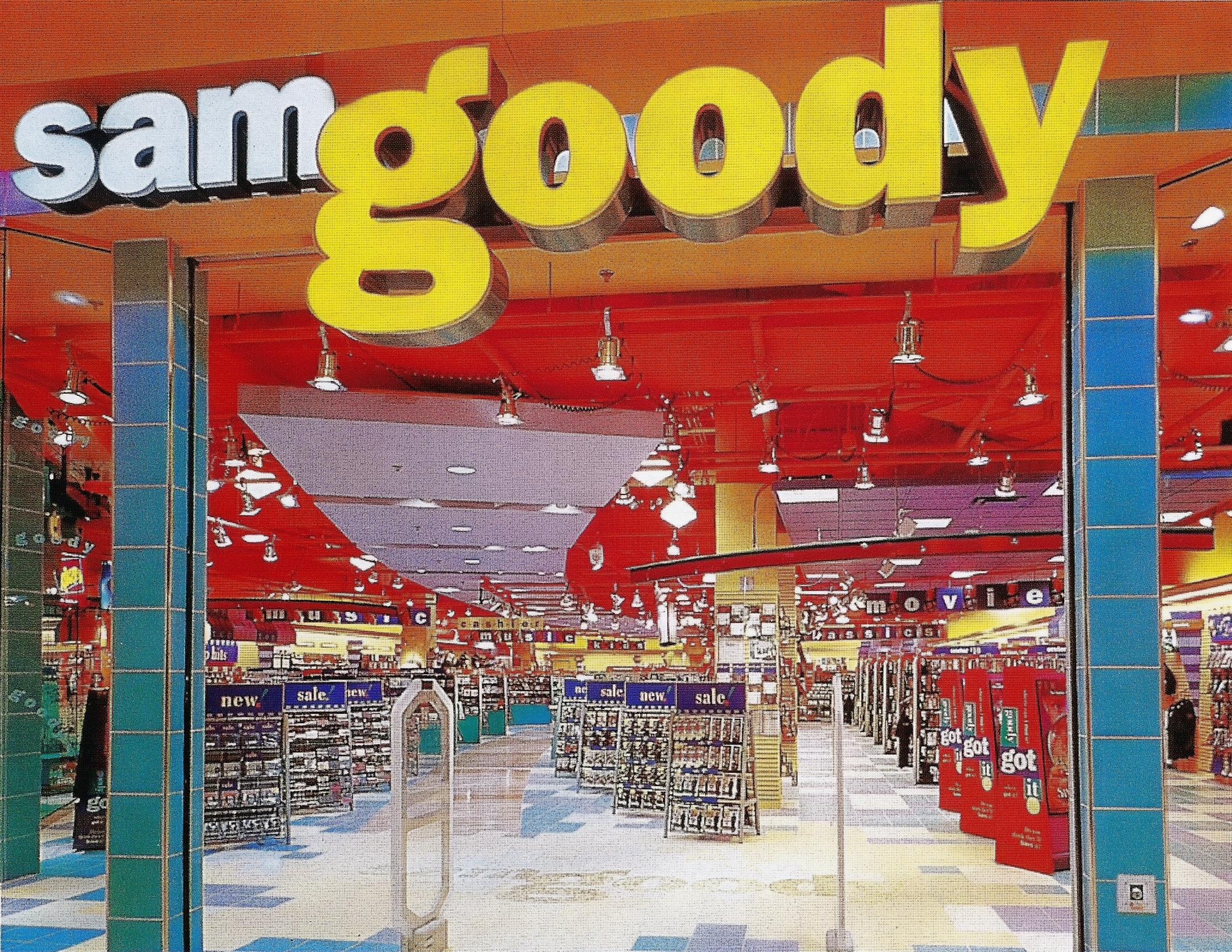 The iconic oversized yellow logo and neon lit entrance of a 90s shopping mall Sam Goody, packed to the ceiling with cassette tapes, CD jewel cases, and towering music promo displays.