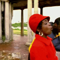 An animated GIF of Lauryn Hill wearing a bright red hat and coat, singing soulfully under stone pillars in The Fugees' "Killing Me Softly" music video.