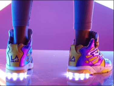 A pair of high-top L.A. Gear Lights sneakers with battery-powered LEDs glowing brightly in the heels, accompanied by black skate shoes.