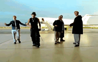AJ McLean in a black fedora and sleeveless shirt gesturing toward the camera, flanked by members of the Backstreet Boys dancing on an airport tarmac with a jet in the background from 'I Want It That Way.'