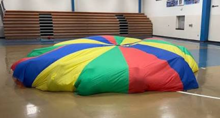 A rainbow gym class parachute billowing up with air on a polished basketball court floor, capturing the single greatest collective moment of pure 90s elementary school joy.