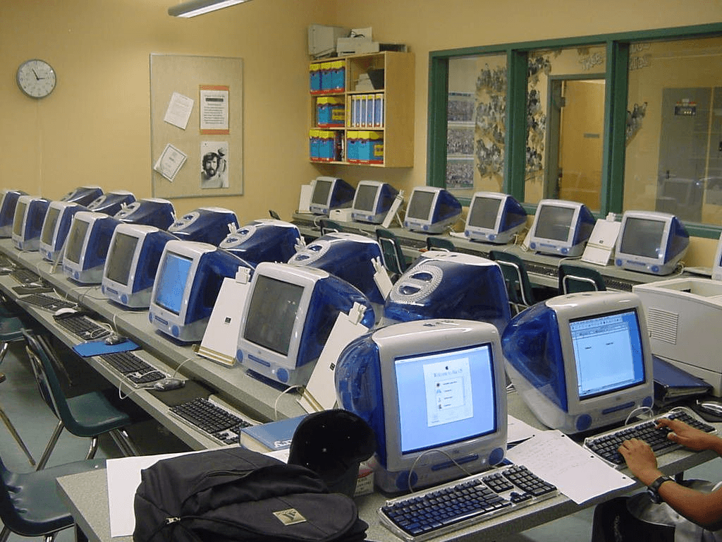 A 90s school computer lab packed with rows of translucent Bondi Blue Apple iMac G3 computers, fully prepped for Oregon Trail runs and Mavis Beacon typing tests.
