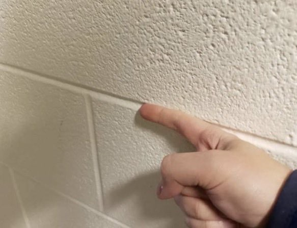 hand tracing a finger along the groove of a painted white cinderblock hallway wall, a universal idle habit for kids walking in line to lunch.