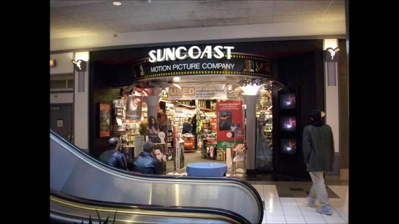 A Suncoast Motion Picture Company mall store entrance surrounded by film-strip decorative trim, wall-mounted preview monitors, and stacks of VHS tapes and early DVDs.