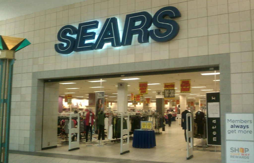 A retro indoor mall storefront shot of Sears, featuring its classic blue block-letter signage above wide open entrance gates lined with security towers.