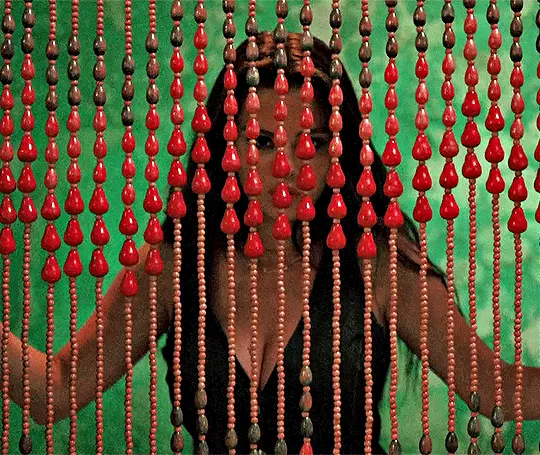 An animated GIF showing a woman in a black dress peering through hanging strands of bright red teardrop-shaped beaded curtains against a green background.