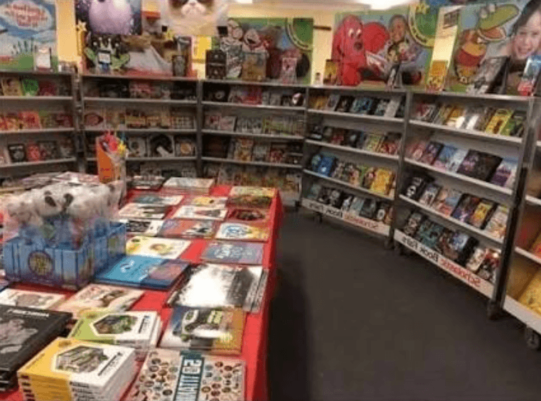 A nostalgic walk through a 90s elementary school library transformed by the Scholastic Book Fair, complete with metal display racks overflowing with paperbacks, posters, and novelty erasers.
