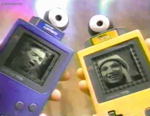 An animated GIF from a retro Game Boy Camera TV commercial showing hands holding purple and yellow Game Boys, each equipped with eyeball-shaped camera cartridges displaying grainy 4-bit pixelated photos on screen.