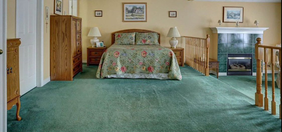 A spacious 90s-style bedroom featuring seafoam green wall-to-wall carpeting, honey oak furniture, a floral quilt, and a tiled gas fireplace.