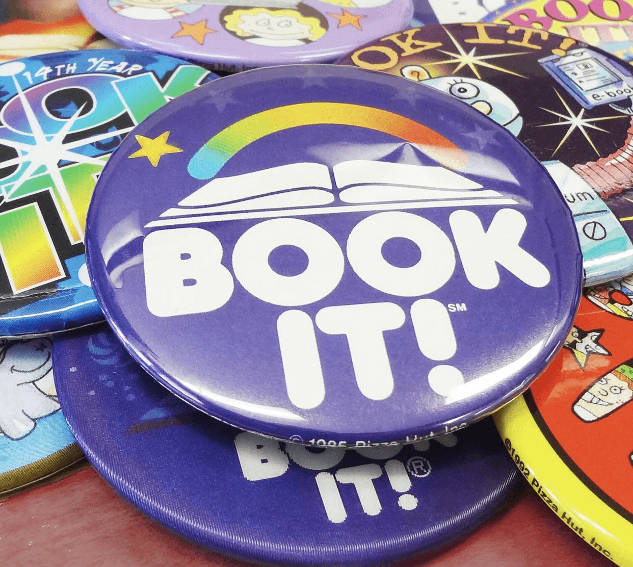 A close-up pile of classic plastic Pizza Hut BOOK IT! pinback buttons, featuring the iconic purple rainbow-and-open-book logo that earned kids free personal pan pizzas.