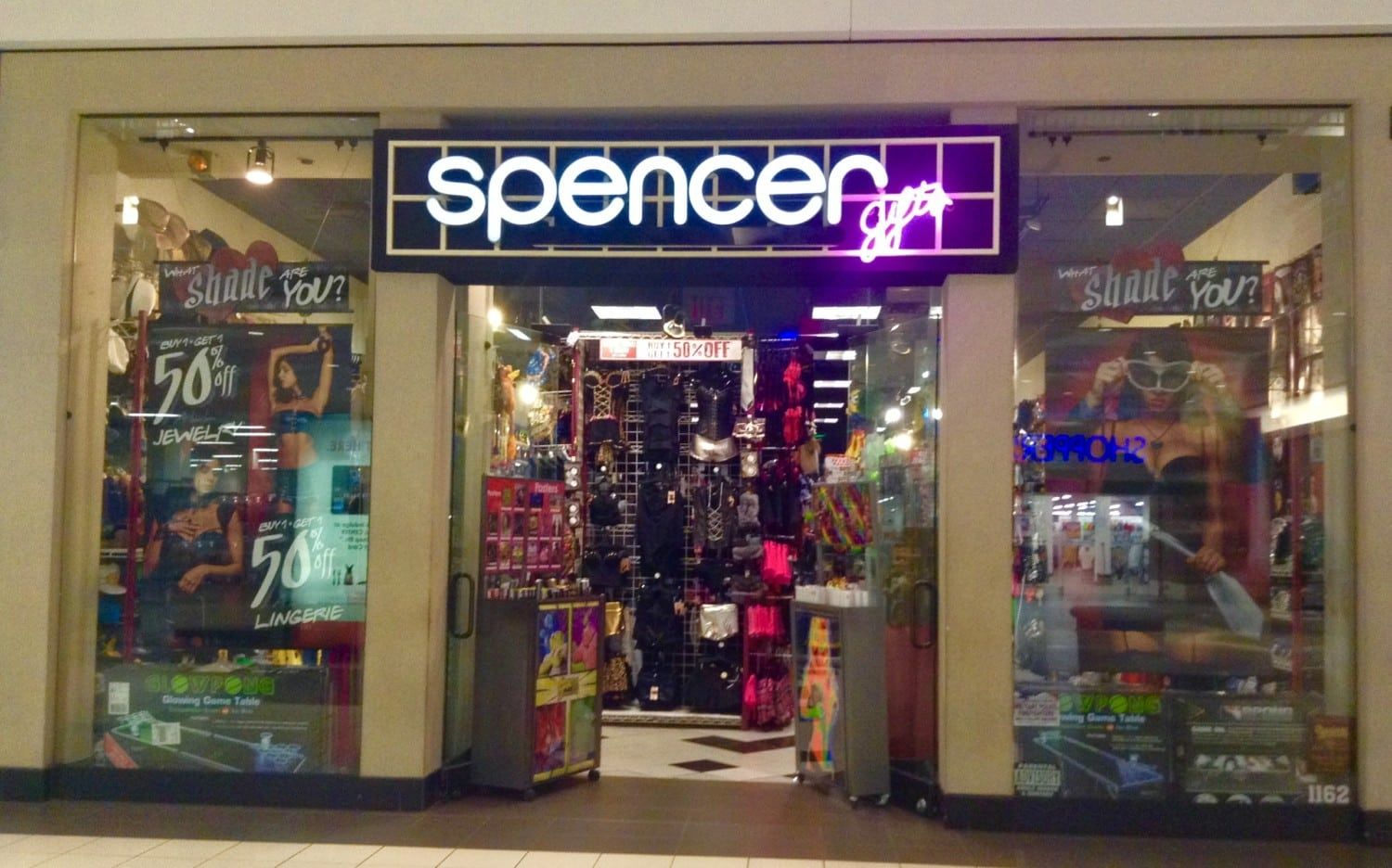 An indoor mall shot of Spencer Gifts, featuring its glowing purple neon sign, dark interior entryway, and racks of novelty t-shirts and pop-culture posters.