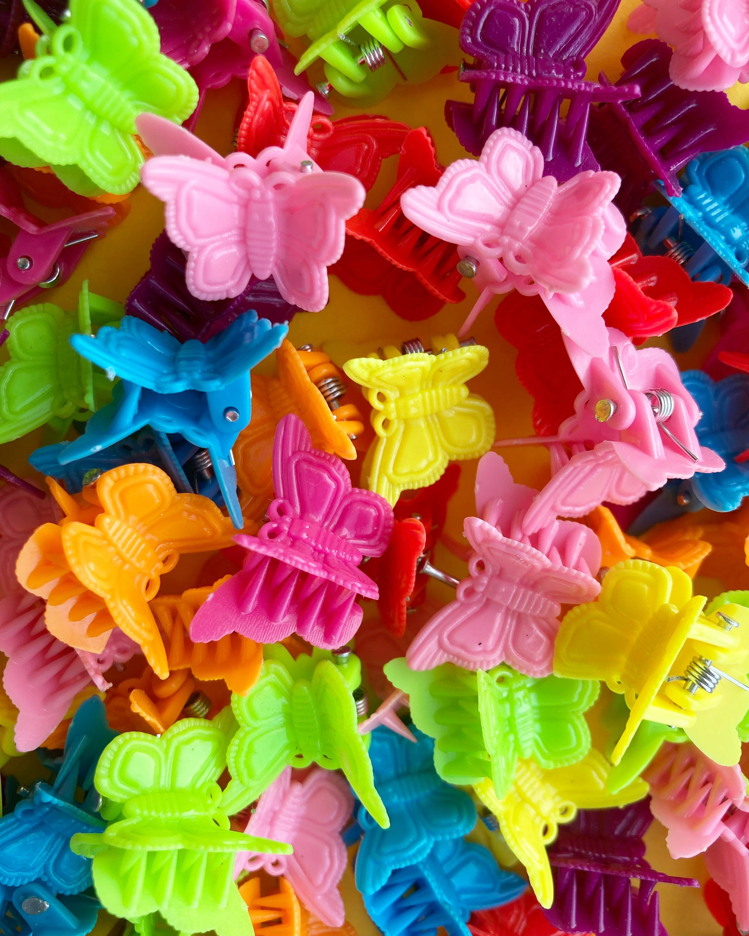 A chaotic, colorful pile of 90s plastic mini butterfly hair clips in neon pink, lime green, bright yellow, and sky blue—the ultimate Y2K hair accessory.