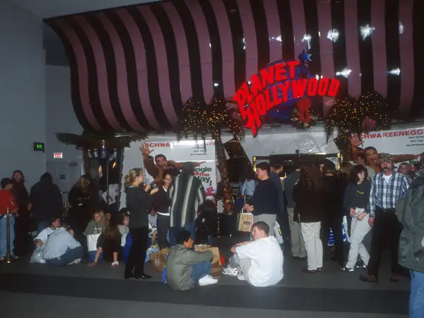A dark interior shot of a 1990s Planet Hollywood restaurant entrance, featuring its marquee sign framed by fake palm trees and movie memorabilia.