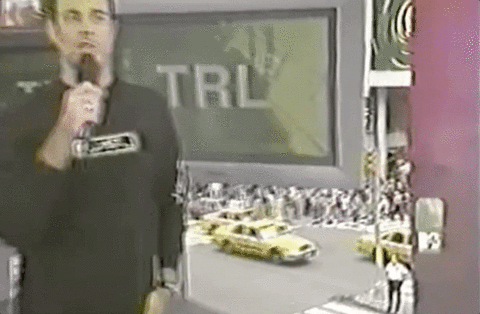 An animated GIF of Carson Daly holding a microphone outside MTV's glass Times Square studio while hosting Total Request Live (TRL) above passing yellow taxis.