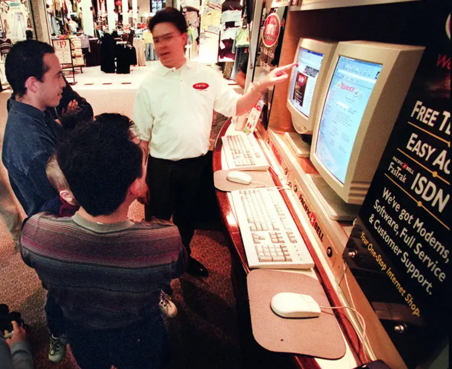 A photo of people gathered around beige CRT computer monitors in an electronics store during the 1990s, with a store clerk pointing at a web page on screen next to ISDN marketing signs.