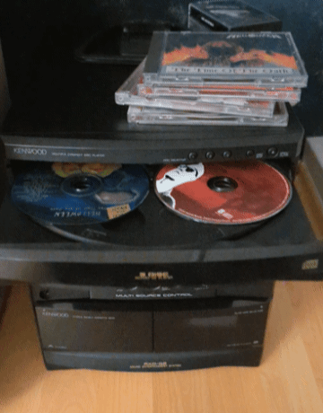 An animated GIF showing an open multi-disc tray on a black Kenwood home stereo system spinning two CDs around, beneath a stack of metal jewel cases including Helloween's The Time of the Oath.