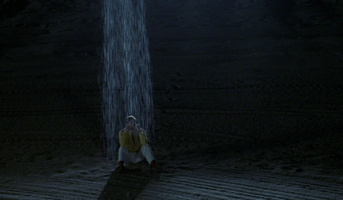 An animated GIF from The Truman Show featuring Truman Burbank (Jim Carrey) sitting huddled on a dark beach as a single, unnaturally localized spotlight rainstorm pours water exclusively down on top of his head.