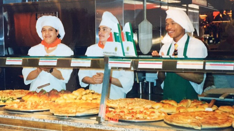 A busy 90s mall food court Sbarro counter with three chefs wearing white hats behind a glass display packed with giant New York-style pizza pies, strombolis, and garlic knots.