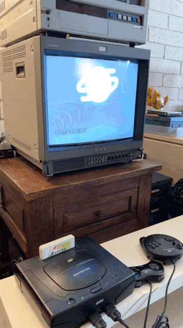 An animated GIF showing a black Sega Saturn console equipped with an Action Replay cartridge powering on, revealing its glowing, metallic orb boot animation on a professional Sony Trinitron CRT monitor.