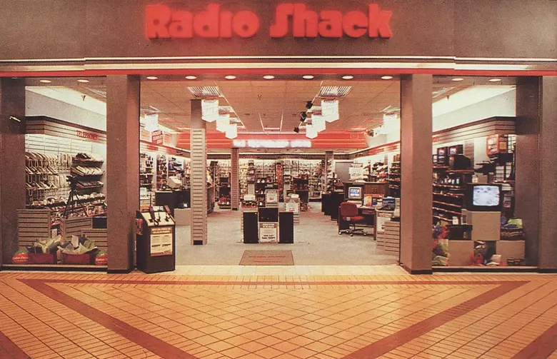 A 90s mall RadioShack entrance glowing with warm red signage, showing off rows of corded landline phones, CRT televisions, and aisles stocked with obscure electronic components.
