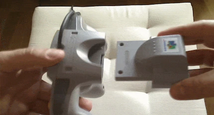 An animated GIF demonstrating hands sliding a grey Nintendo 64 Rumble Pak accessory into the bottom expansion slot of an N64 controller—the force-feedback revolution that drained AA batteries and rattled hands nationwide.