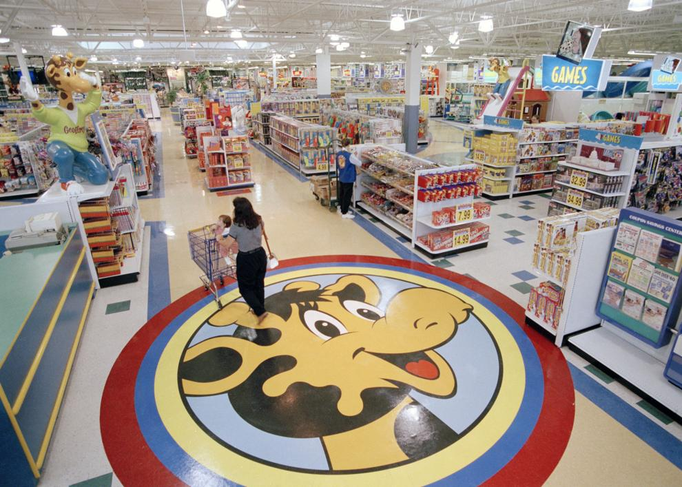 An overhead view inside a packed 90s Toys "R" Us store, showing customers pushing shopping carts over a massive circular floor decal of Geoffrey the Giraffe surrounded by endless aisles of board games and action figures.