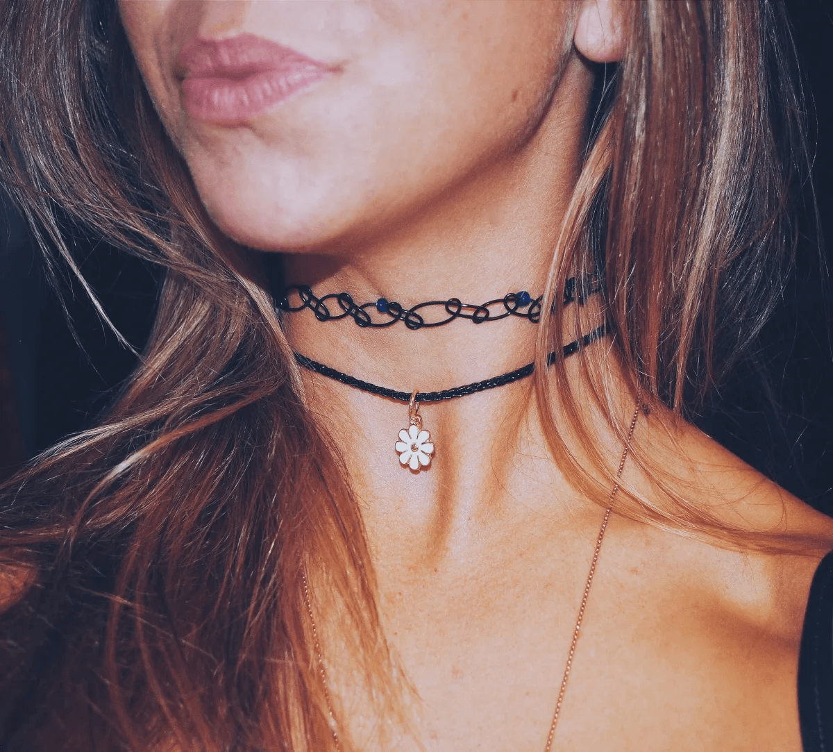 A close-up shot of 90s neckwear trends, showing layered black elastic stretch tattoo chokers paired with a silver daisy pendant necklace.