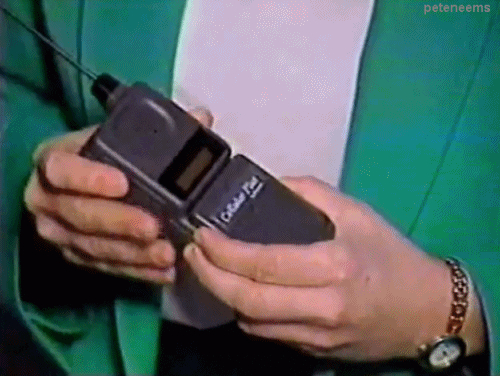 90s TV tech segment showing hands opening up a chunky, black cellular flip phone with a pop-up whip antenna.