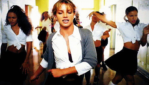 Britney Spears wearing braided pigtails, a tied-up white button-down shirt, and a cardigan, dancing down a school hallway surrounded by backup dancers in her 1998 '...Baby One More Time' music video.