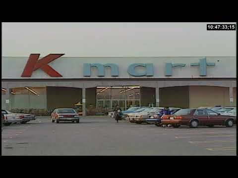 A wide outdoor shot of a classic 1990s Kmart anchor store exterior, featuring the signature red 'K' logo and a parking lot filled with period-accurate cars.