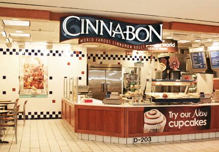 A retro Cinnabon mall kiosk with tiled counter fronts, stainless steel warming racks full of frosted cinnamon rolls, and overhead menu boards beckoning shoppers with irresistible smells.