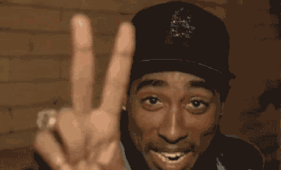 An animated GIF of Tupac Shakur smiling warmly at the camera, raising two fingers to flash a peace sign while wearing a black fitted cap.